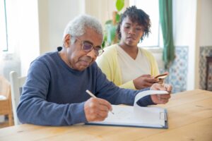 Home A senior man writing on papers with a caregiver nearby indoors in a supportive setting.